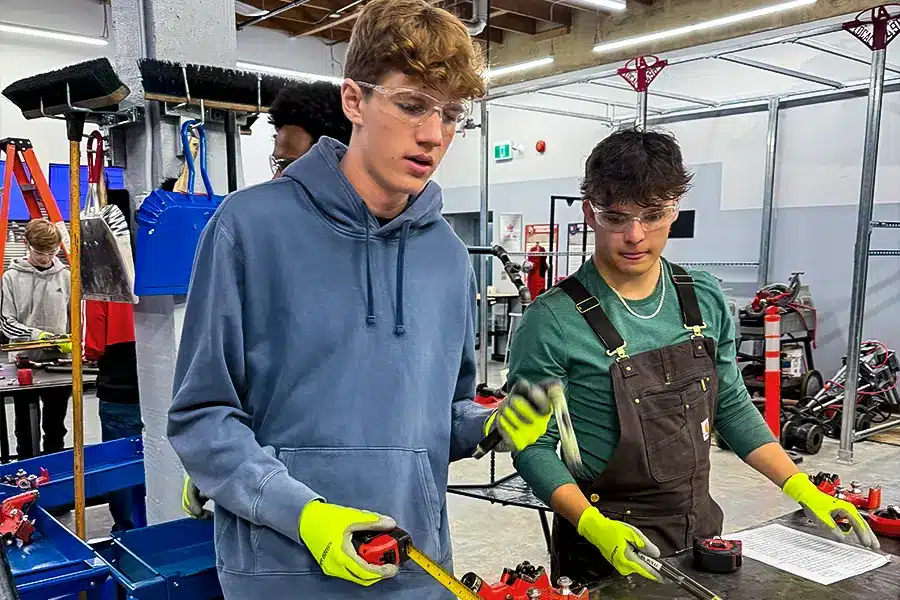 Two Fusion students using a tape measure in a trades shop while working on pipe components.